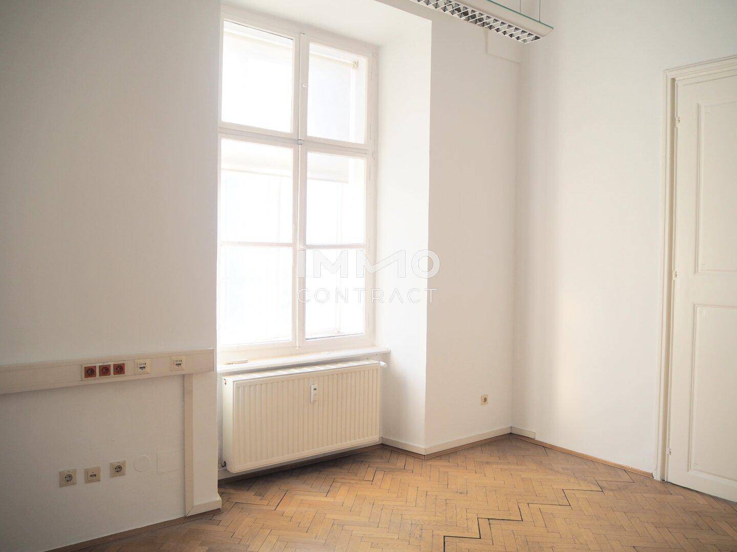 Heller Büroraum mit Fischgrätparkett, hohen Fenstern und Heizkörper unter der Fensterbank, gut belichtet.