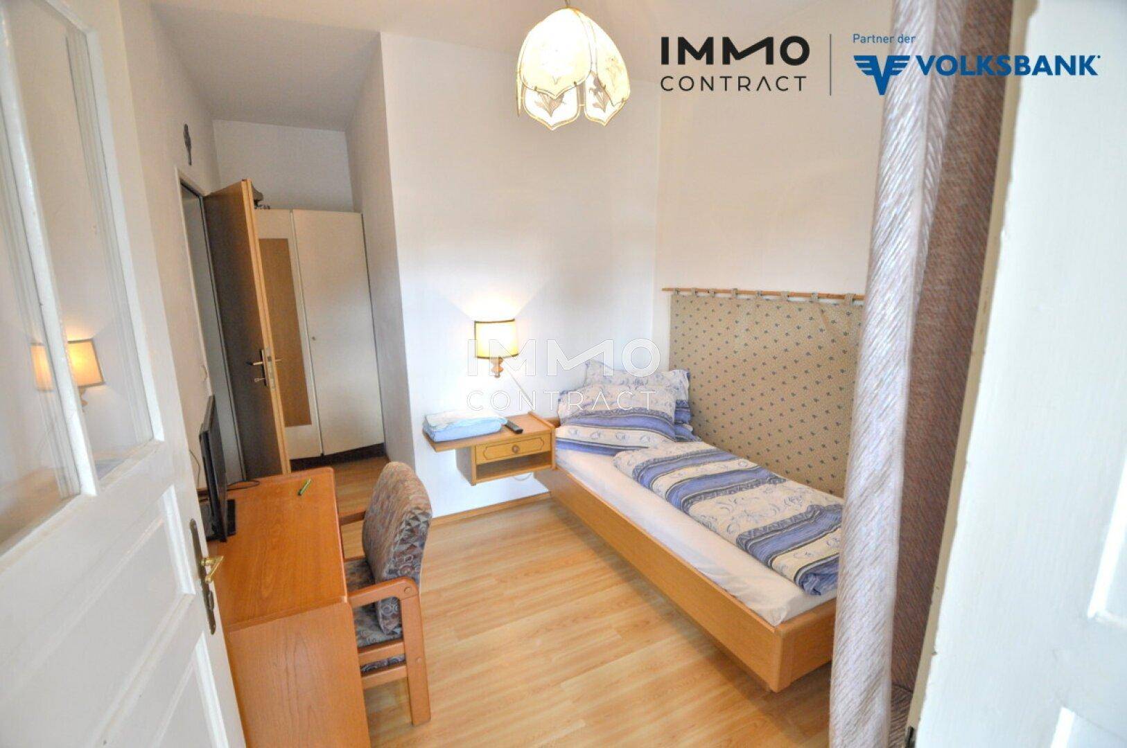 Einfaches Einzelzimmer mit Holzboden, Nachttisch und einem kleinen Schrank.