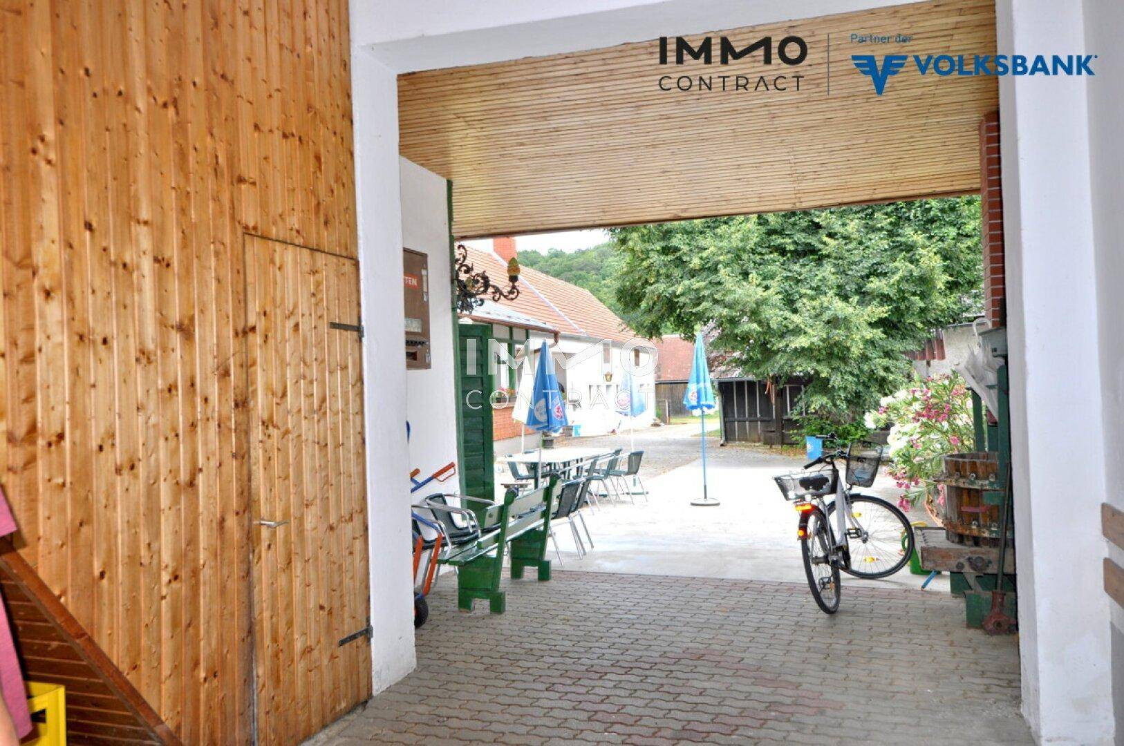 Blick durch einen Holztorbogen auf einen gepflasterten Innenhof mit Sitzgelegenheiten und viel Grün.