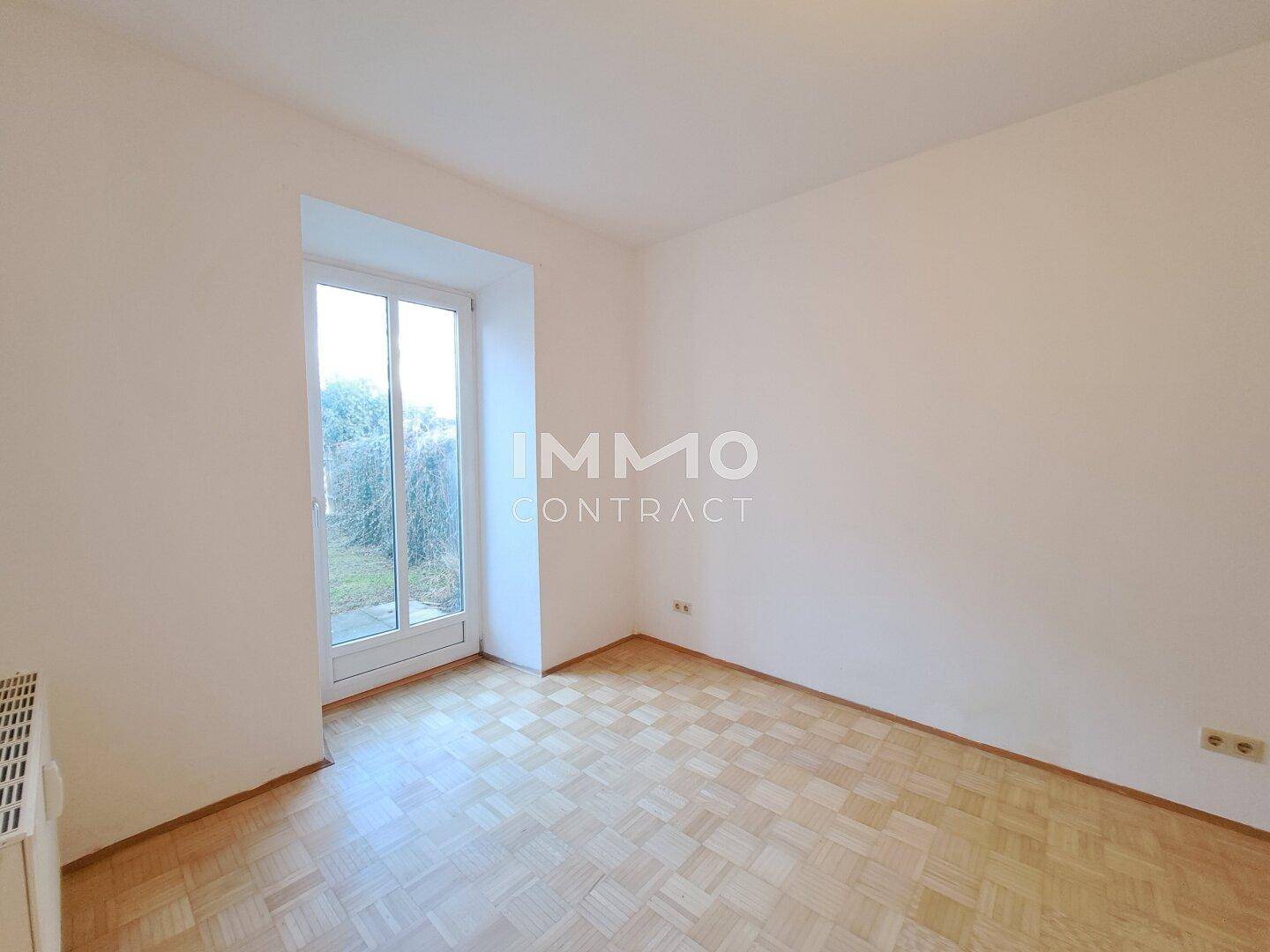 Helles Zimmer mit Parkettboden und großem Fenster mit direktem Blick und Zugang zum Garten.