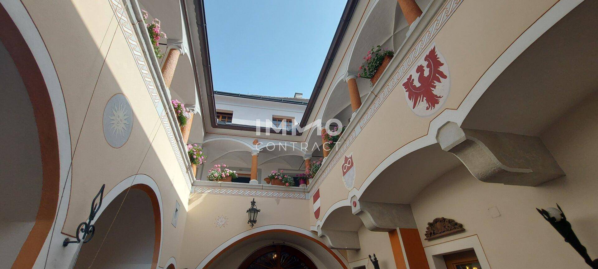 Blick nach oben in einen historischen Innenhof mit Arkaden, verzierten Fassaden und Blumenkästen auf den oberen Etagen.