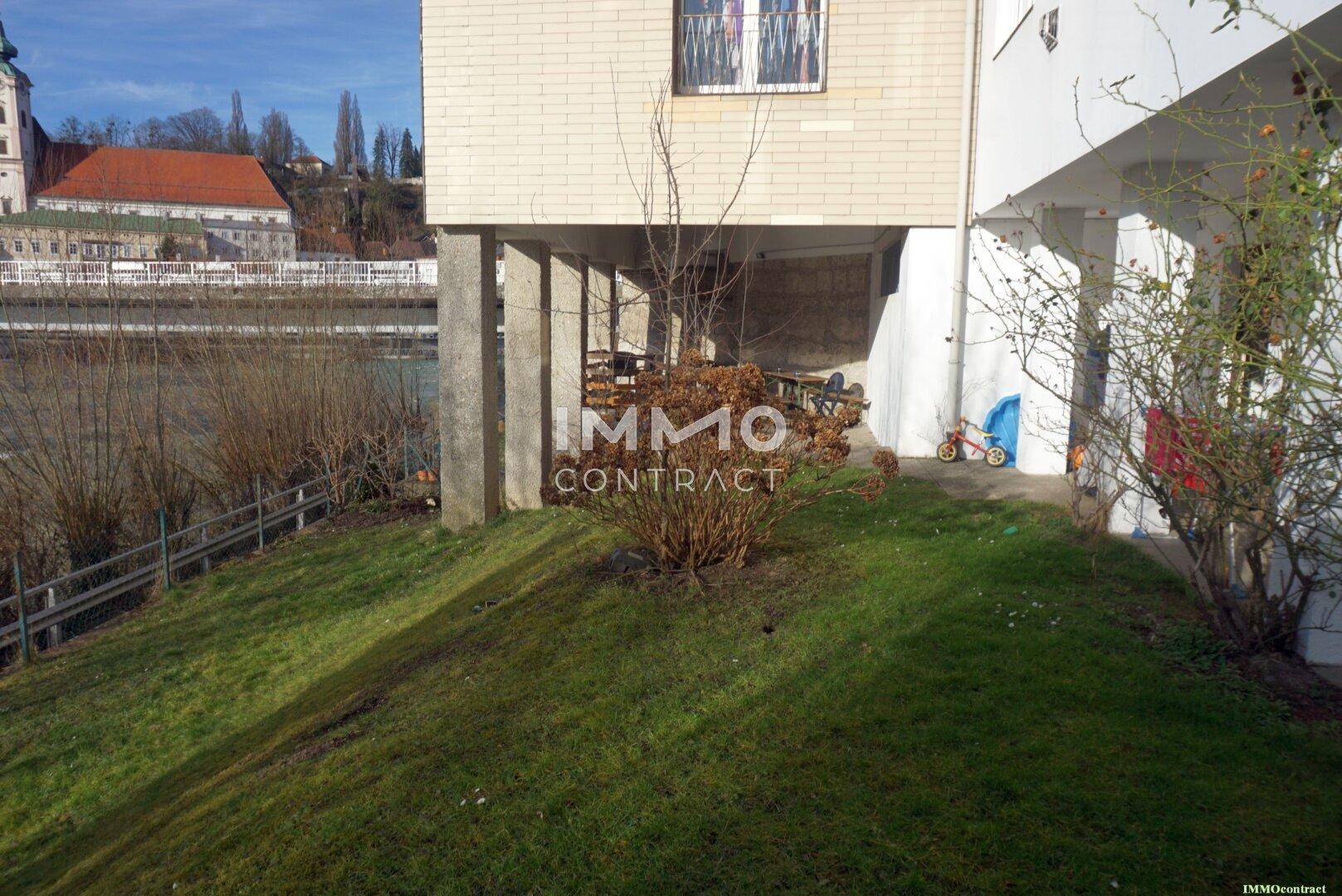 Grünfläche am Flussufer neben einem Gebäude mit Blick auf die gegenüberliegende Stadt und Natur.