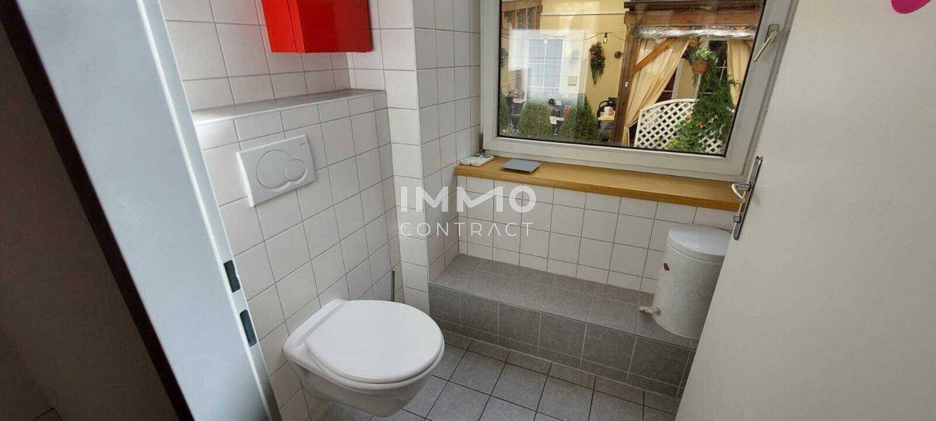 Kleiner Toilettenraum mit weißen Fliesen, Fenster und Blick in einen Innenhof.