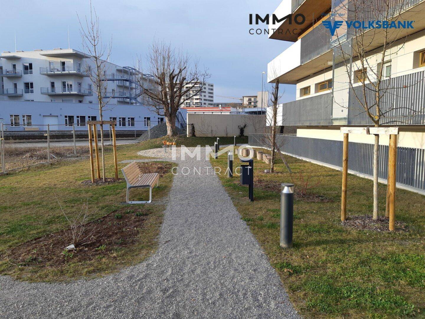 Gemeinschaftlicher Gartenbereich mit Kiesweg, Bänken und jungen Bäumen.