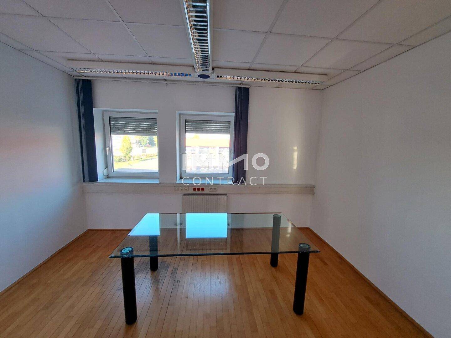 Leeres Büro mit Holzboden, einem Glastisch und Fenstern mit Jalousien.