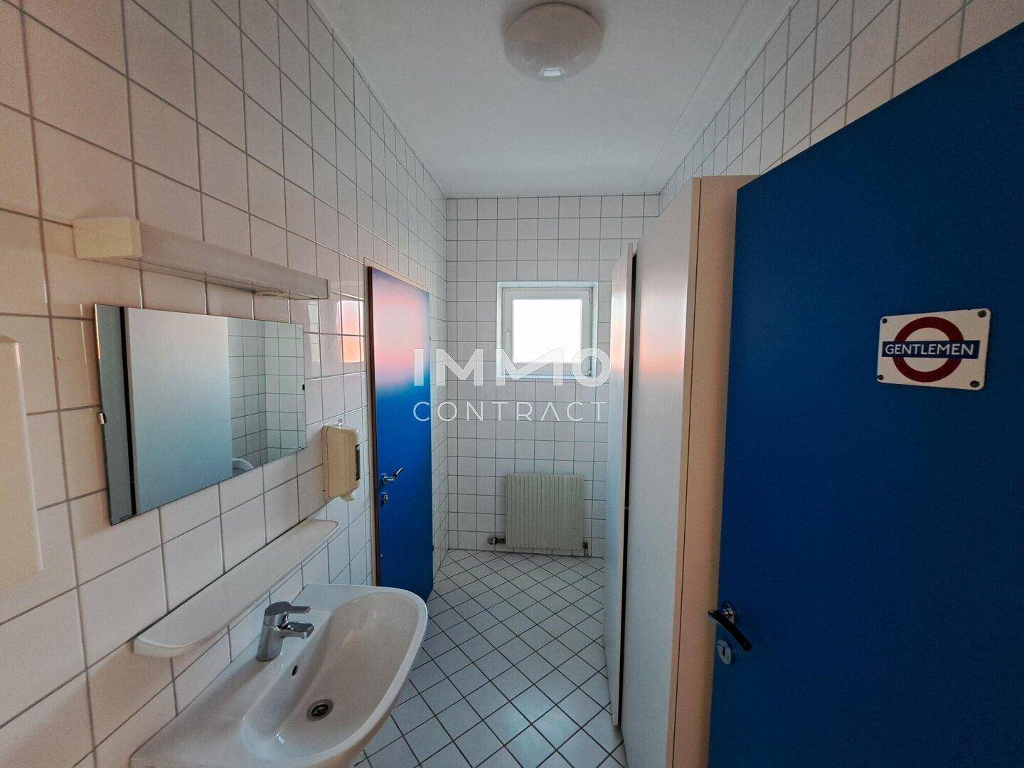 Langer Toilettenbereich mit weiß gefliesten Wänden, Waschbecken und einem Fenster für Tageslicht.