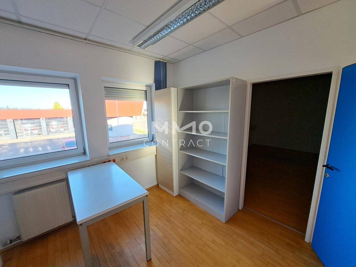 Helles Büro mit Holzboden, einem Tisch und einem Regal, beleuchtet durch Fenster mit Jalousien.