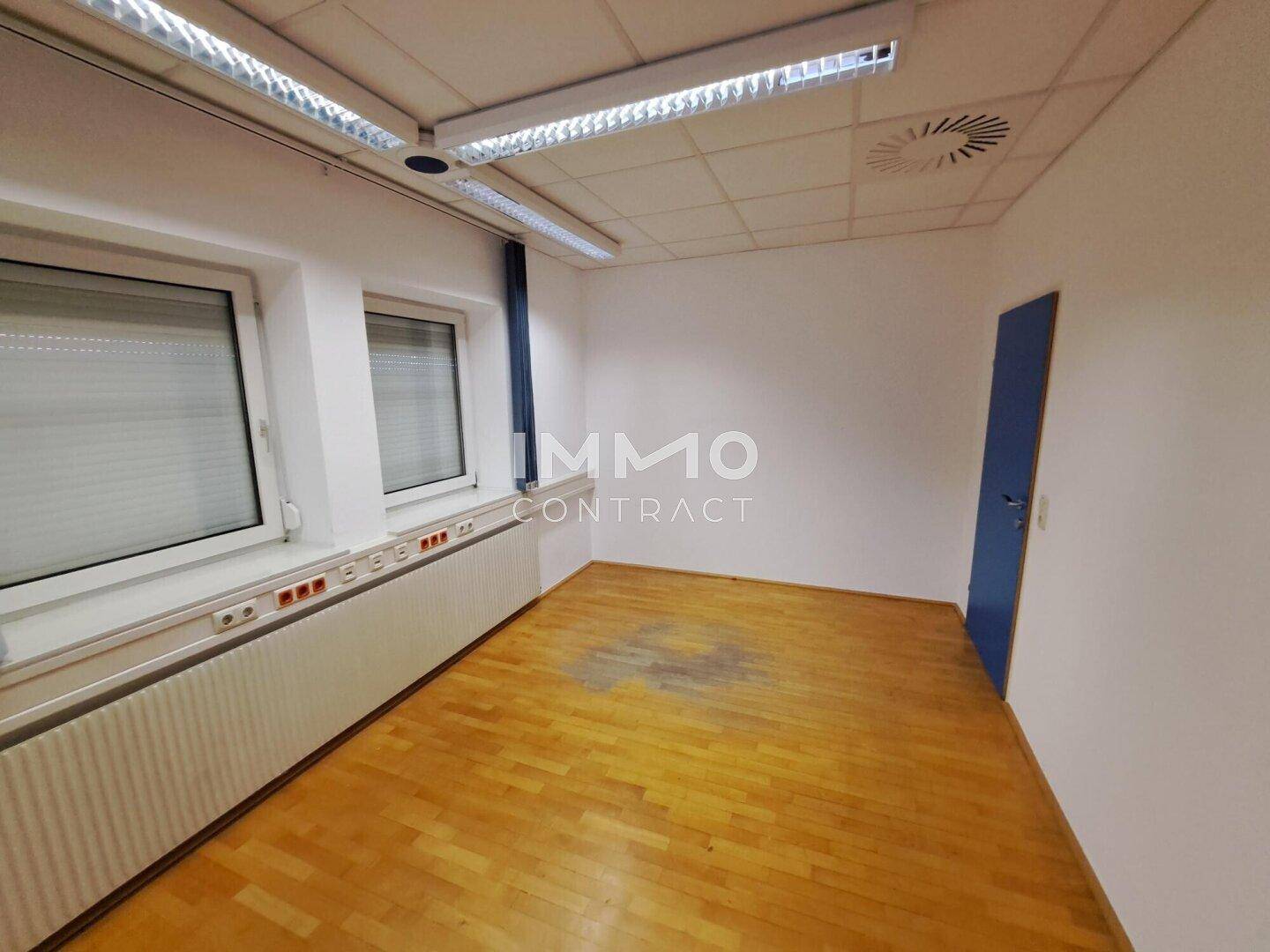 Leeres Büro mit Holzboden, weißen Wänden und Fenstern mit Jalousien.