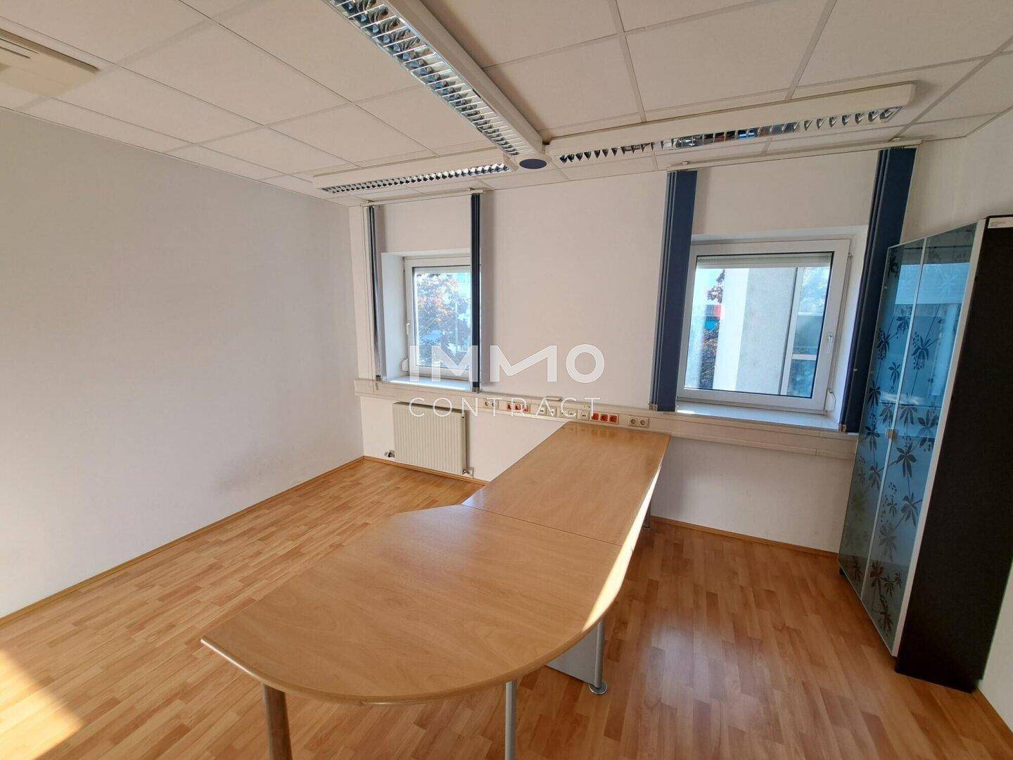 Helles Büro mit Holzboden, einem großen Schreibtisch und Fenstern mit Jalousien.