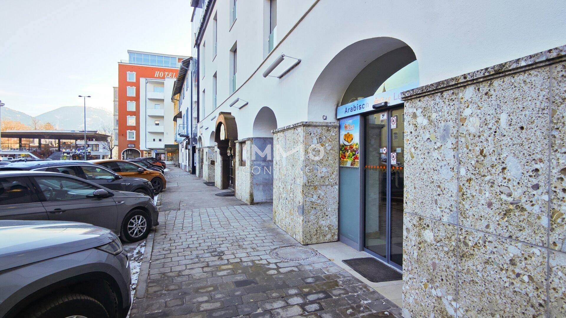 Gebäudefassade mit Arkaden und Natursteinverkleidung entlang einer Straße mit parkenden Fahrzeugen und Bergen im Hintergrund.