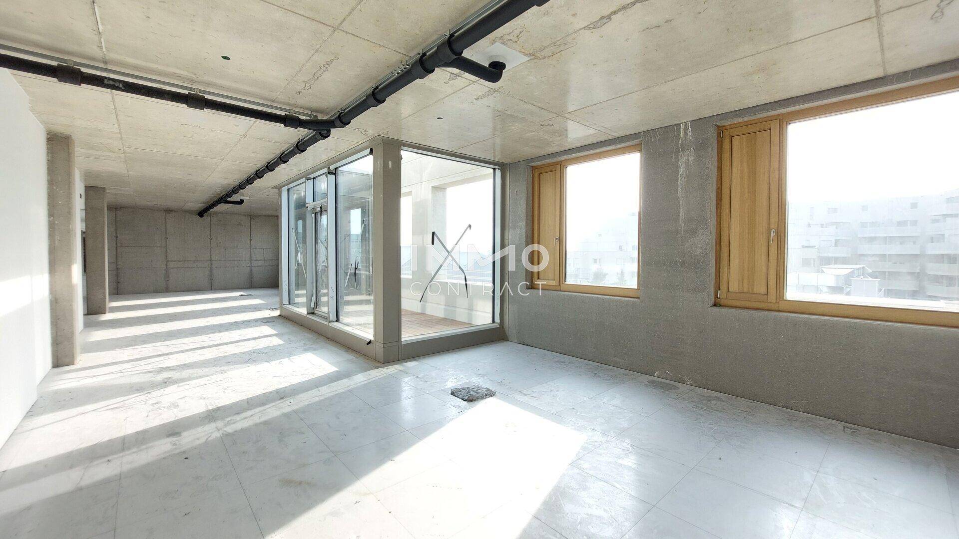 Unfertiger Büroraum mit Betonwänden, sichtbaren Leitungen, Glaskabine und großen Fenstern.