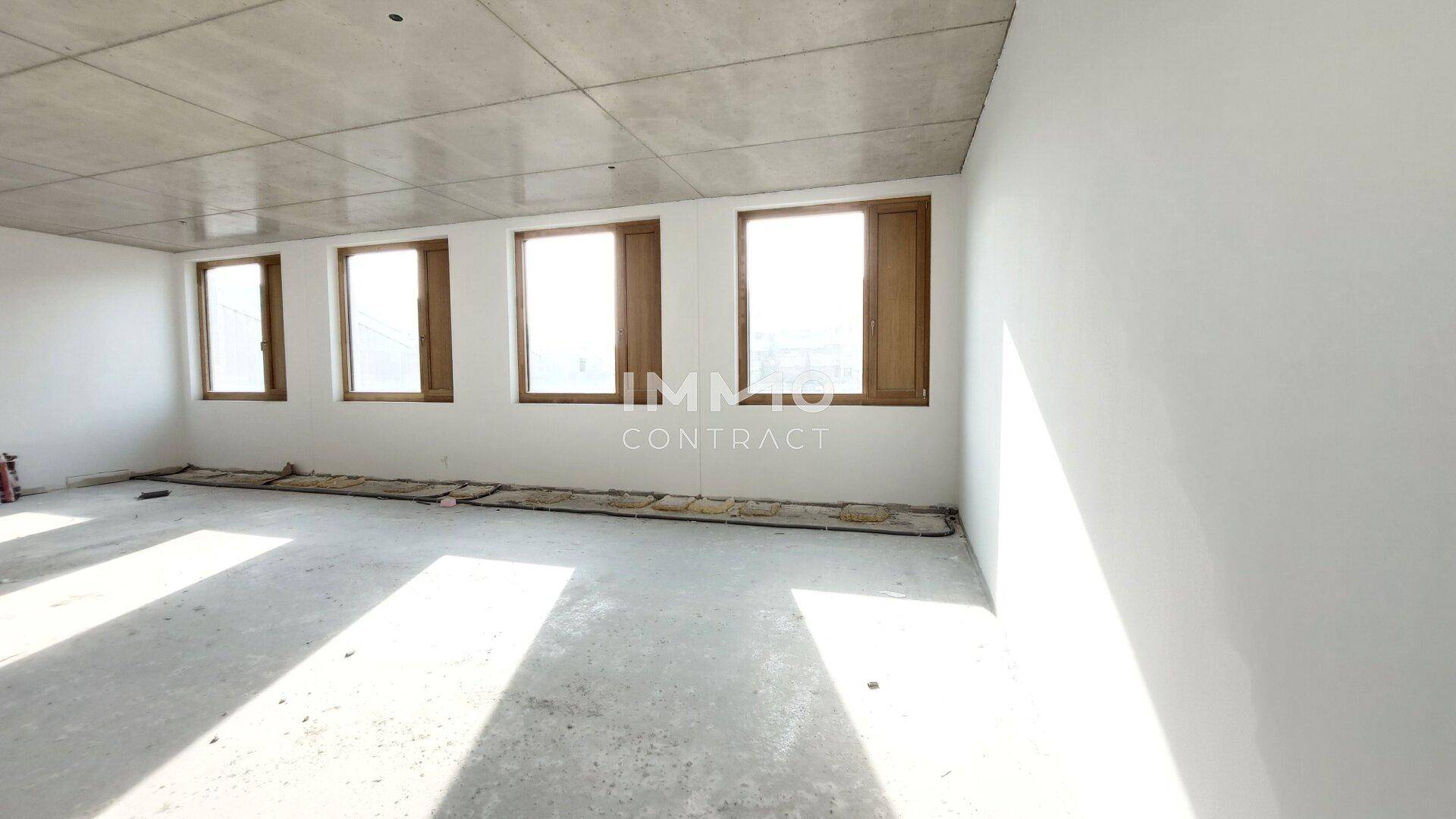 Leerer Büroraum im Rohbau mit weiß gestrichenen Wänden, Betonboden und mehreren Fenstern für viel Tageslicht.