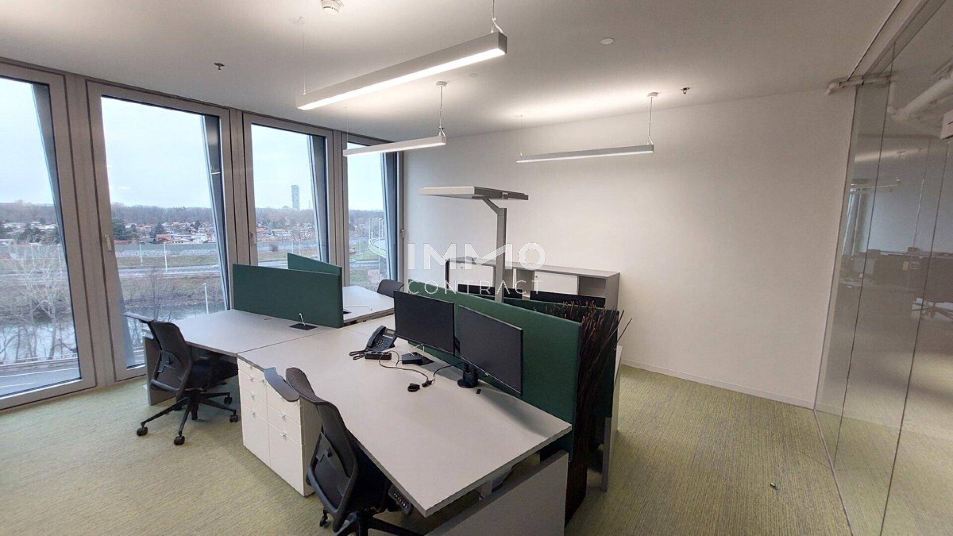 Heller Büroraum mit mehreren Arbeitsplätzen, großen Fenstern und Blick ins Grüne.