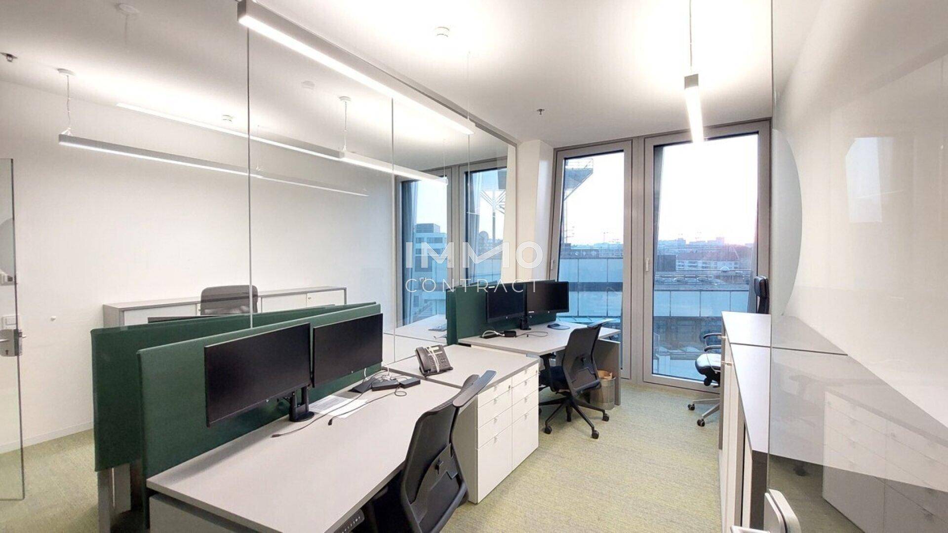 Heller Bürobereich mit mehreren Arbeitsplätzen, grünen Trennwänden und raumhohen Fenstern mit Stadtblick.