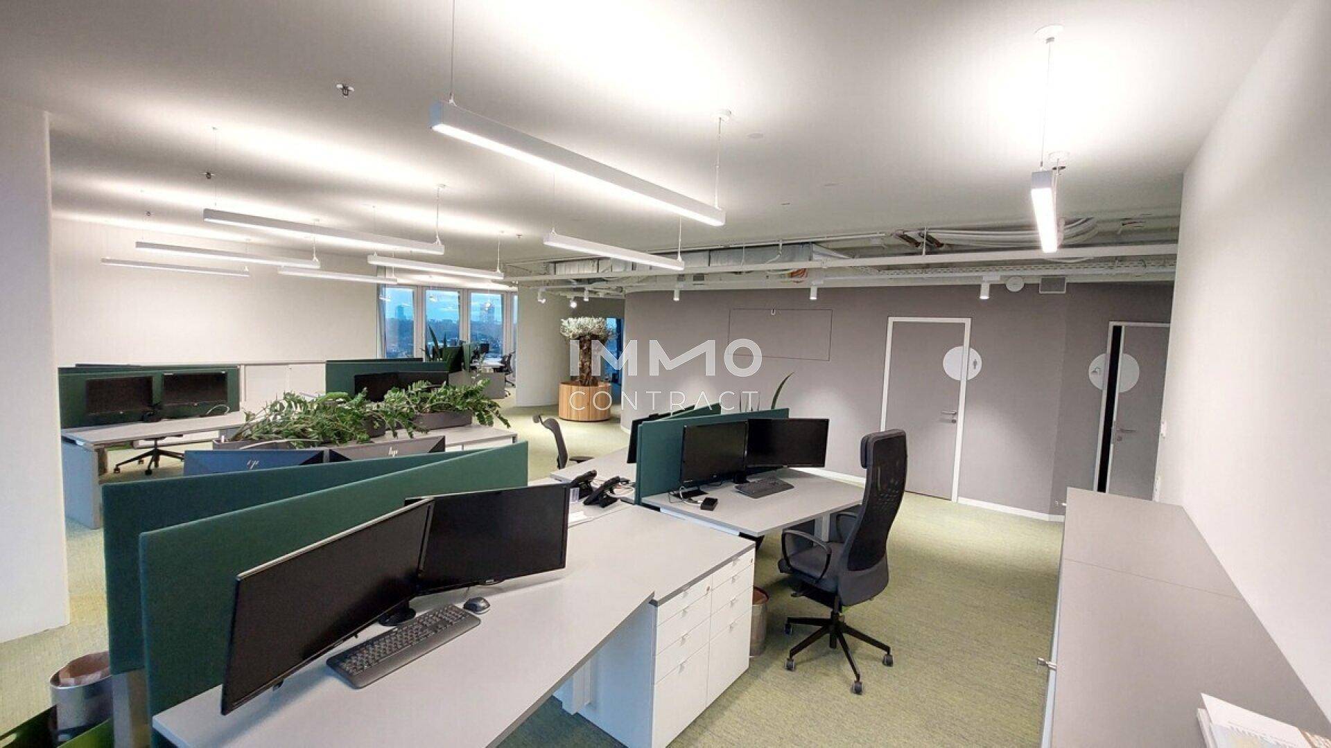 Moderner Bürobereich mit Arbeitsplätzen, grünen Trennwänden und einer grauen Wand mit Türen, sowie sichtbaren Leitungen.