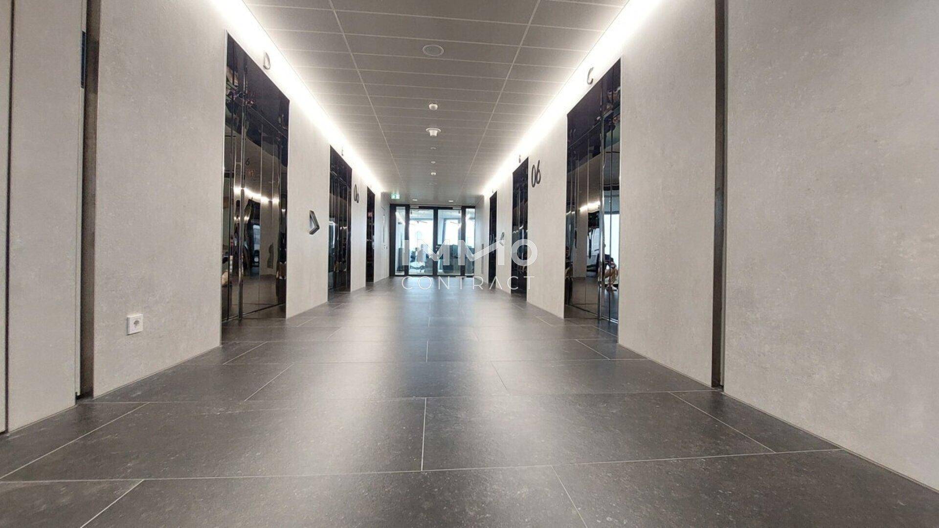 Langer, moderner Flur mit dunklen Fliesen, beleuchteter Decke und Aufzügen auf beiden Seiten.
