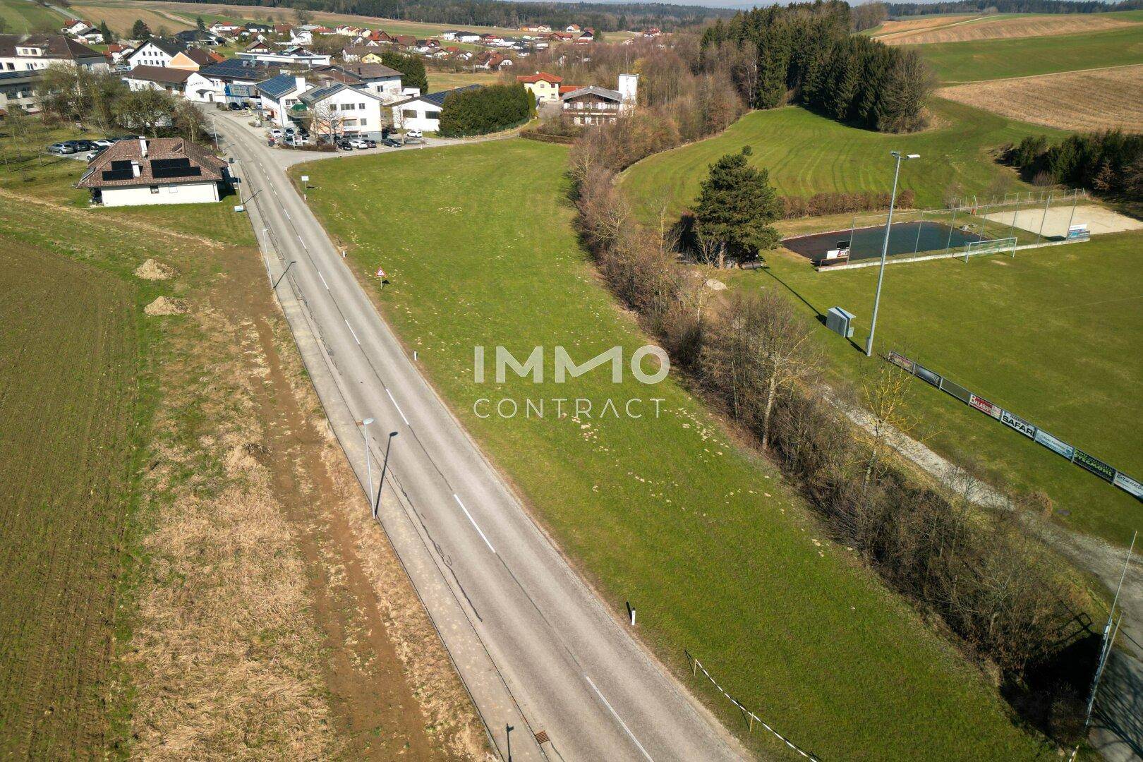 Weitwinkel-Luftaufnahme des Grundstücks mit Straße, Sportplatz und dem dahinterliegenden Dorf unter klarem Himmel.
