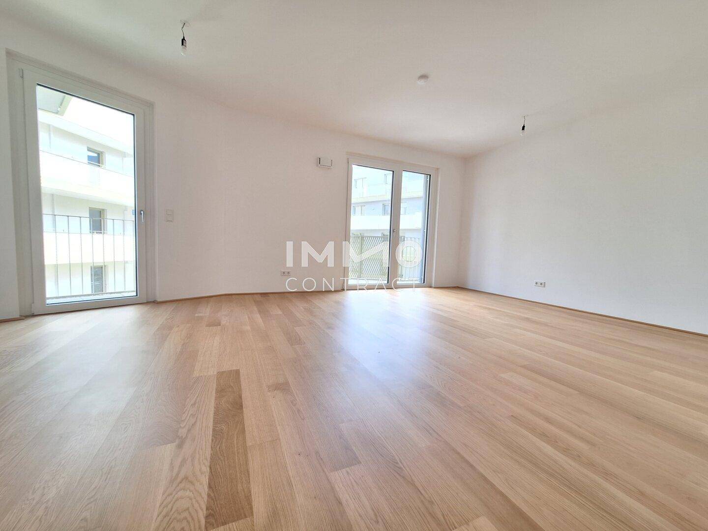 Leeres, lichtdurchflutetes Wohnzimmer mit Parkettboden und großen Fenstern zum Balkon.