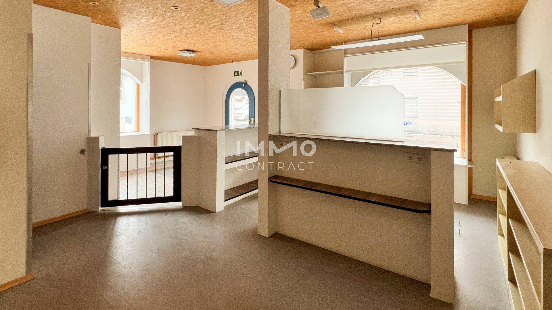 Geräumiger Gewerberaum mit Thekenbereich, großen Fenstern und einer Decke aus Holzspanplatten, offen gestaltet.