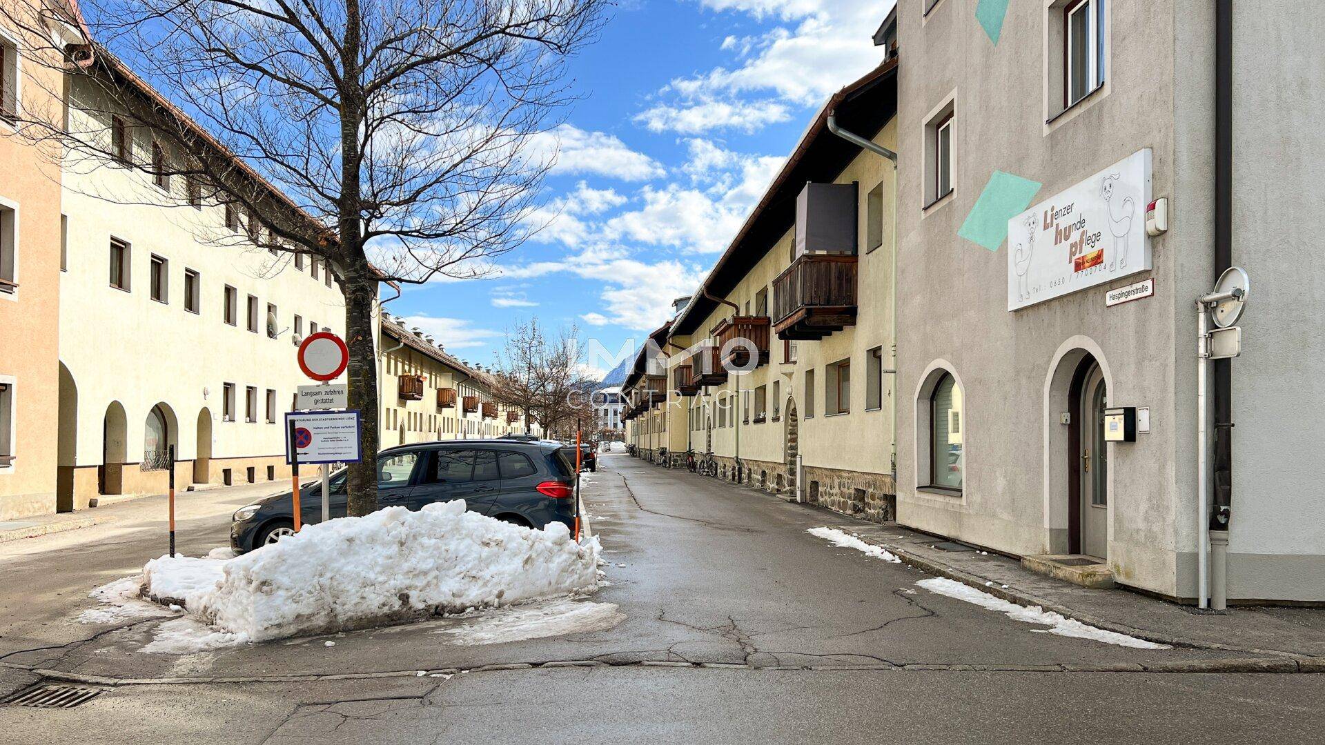 Blick entlang einer verschneiten Straße mit Gebäuden auf beiden Seiten und einem geparkten Auto im Vordergrund.