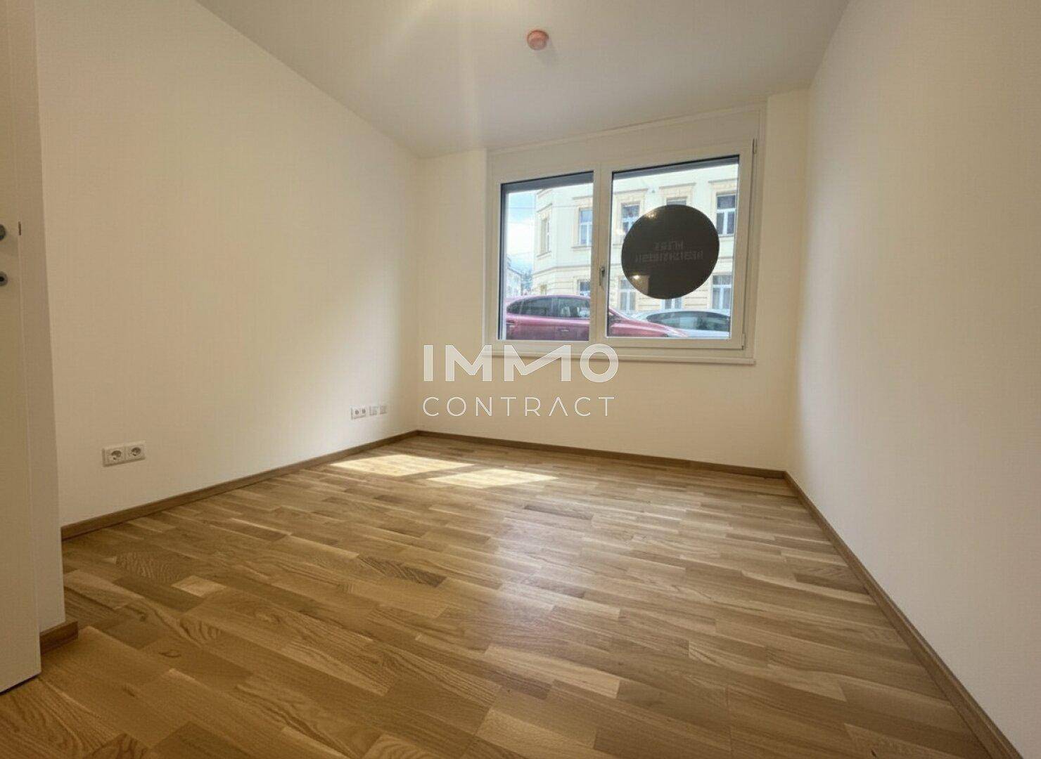 Heller, unmöblierter Raum mit Parkettboden und einem großen Fenster zur Straße hin, das viel Tageslicht bietet.
