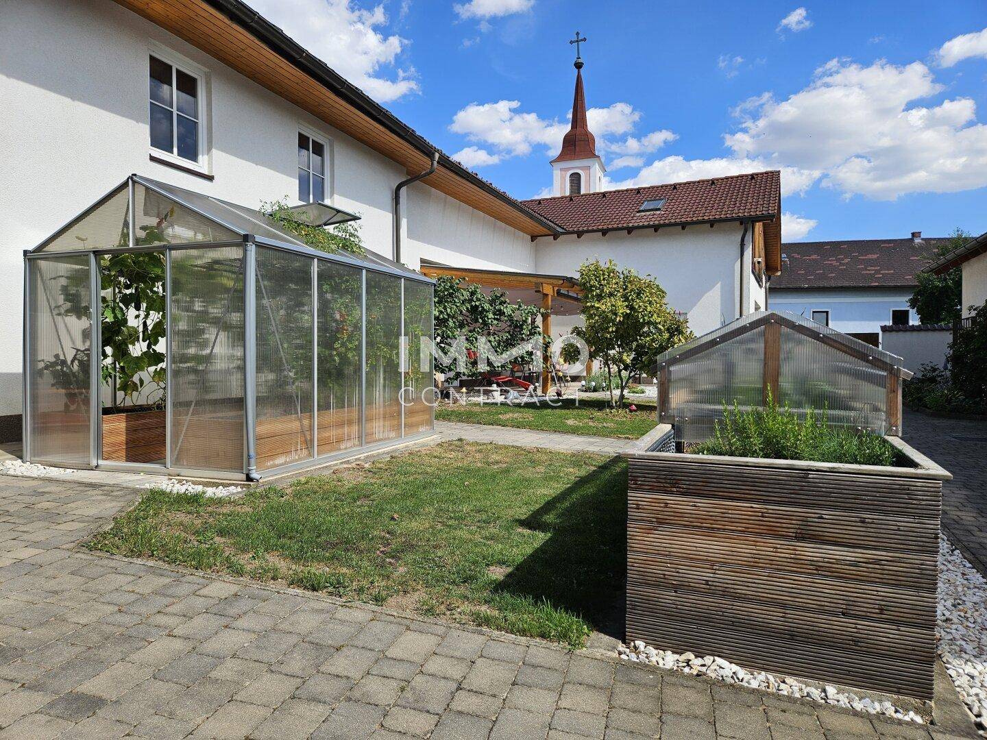 Sonniger Innenhof mit Gewächshaus, Hochbeeten und Blick auf die Kirche im Hintergrund.