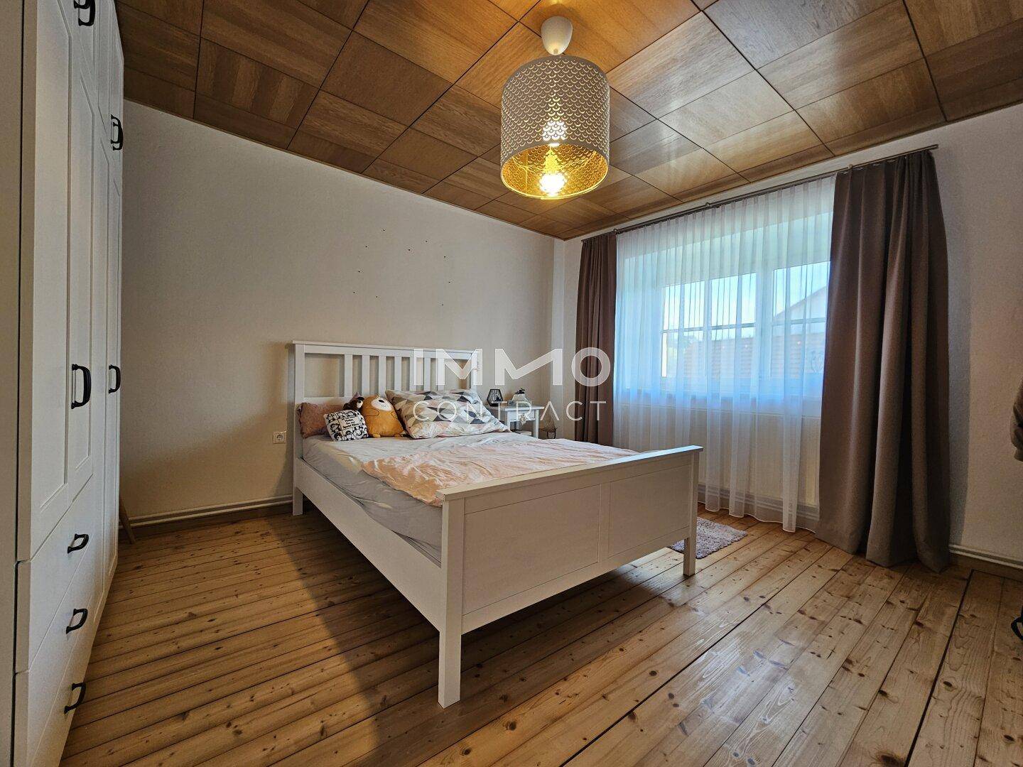 Schlafzimmer mit Holzdielenboden, Holzdecke und einem großen Fenster mit Vorhängen.