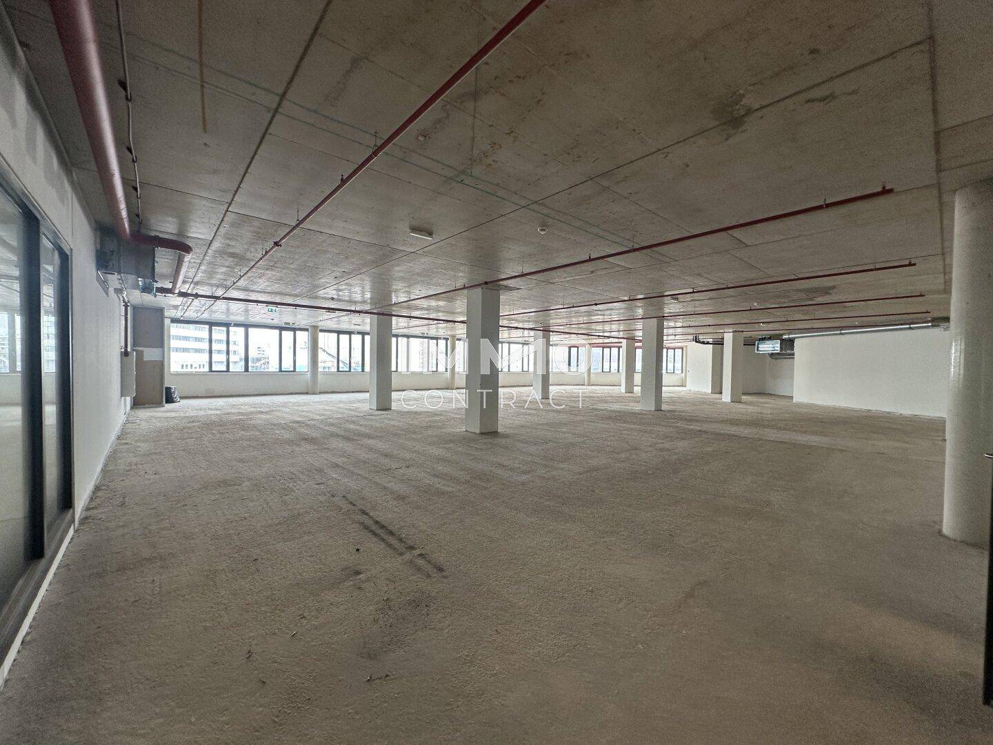 Großer, unfertiger Bürobereich mit Betonboden, sichtbaren Leitungen und großen Fenstern.