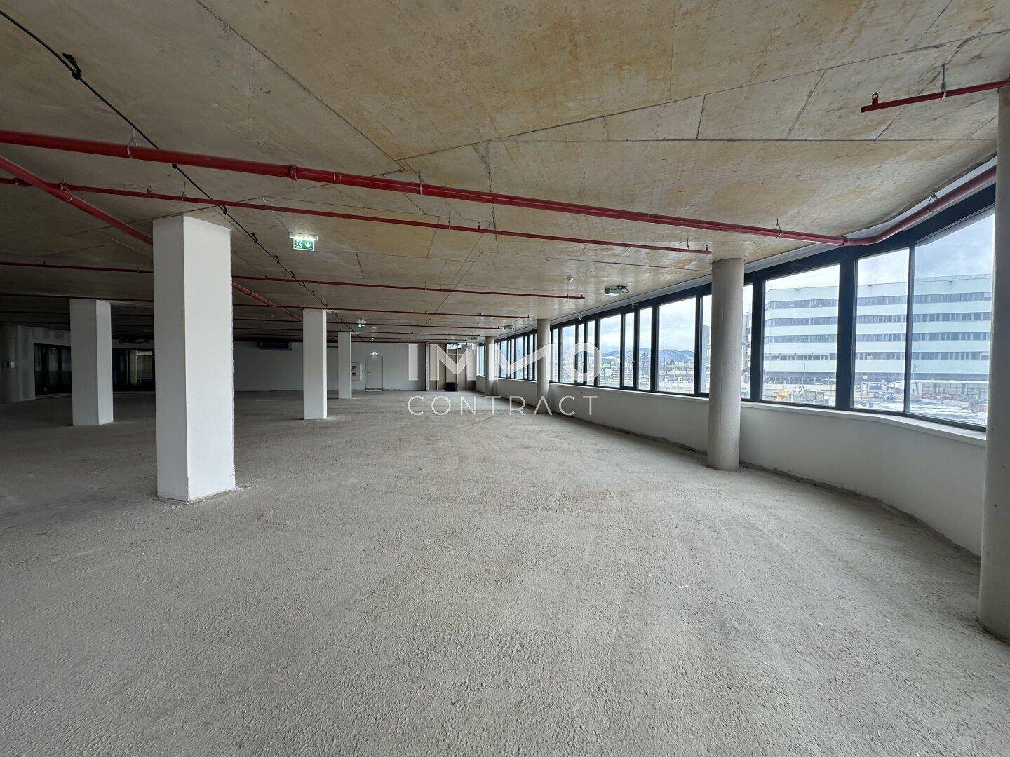 Weitläufiger, unmöblierter Büroraum mit Betonoberflächen und vielen Fenstern für Tageslicht.