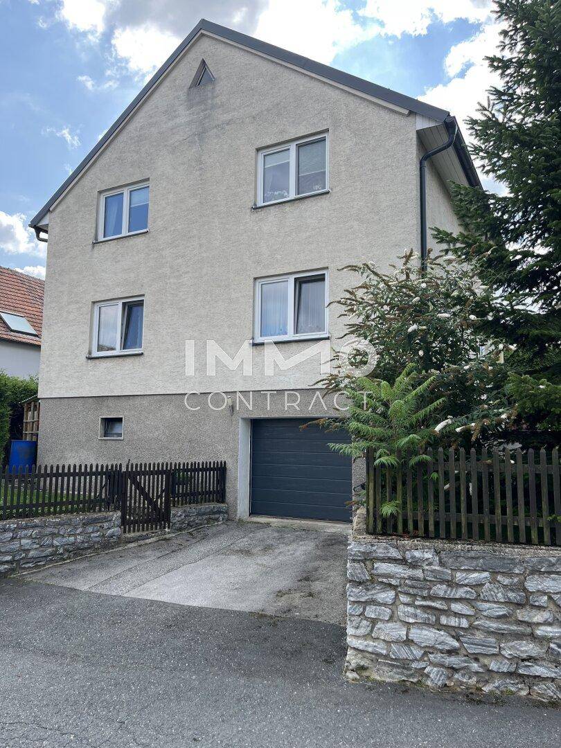Hausfassade mit integrierter Garage und Zufahrt, umgeben von einem Holzzaun und Bäumen.