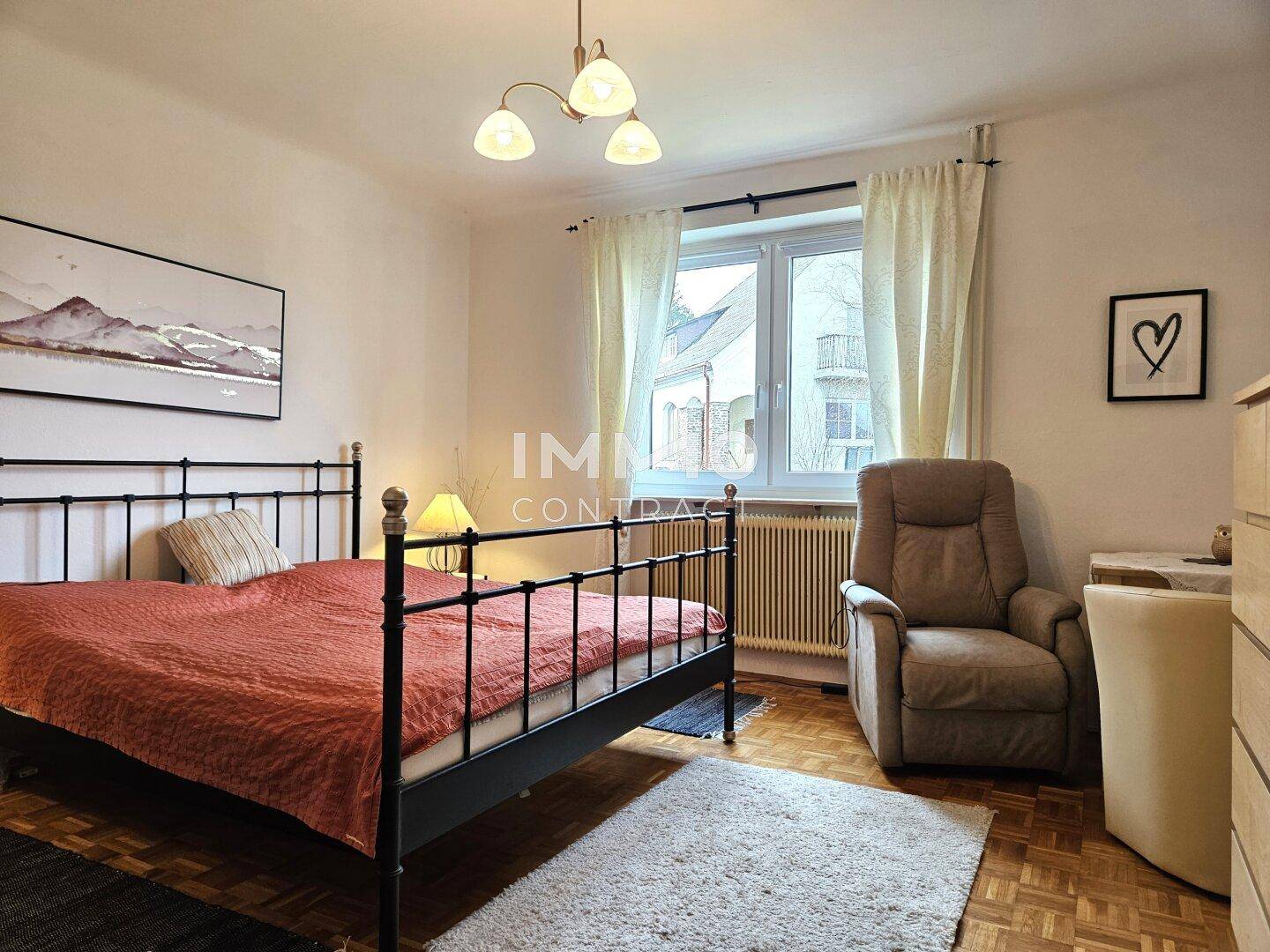 Schlafzimmer mit Parkettboden, Metallbett und einem bequemen Sessel am Fenster.