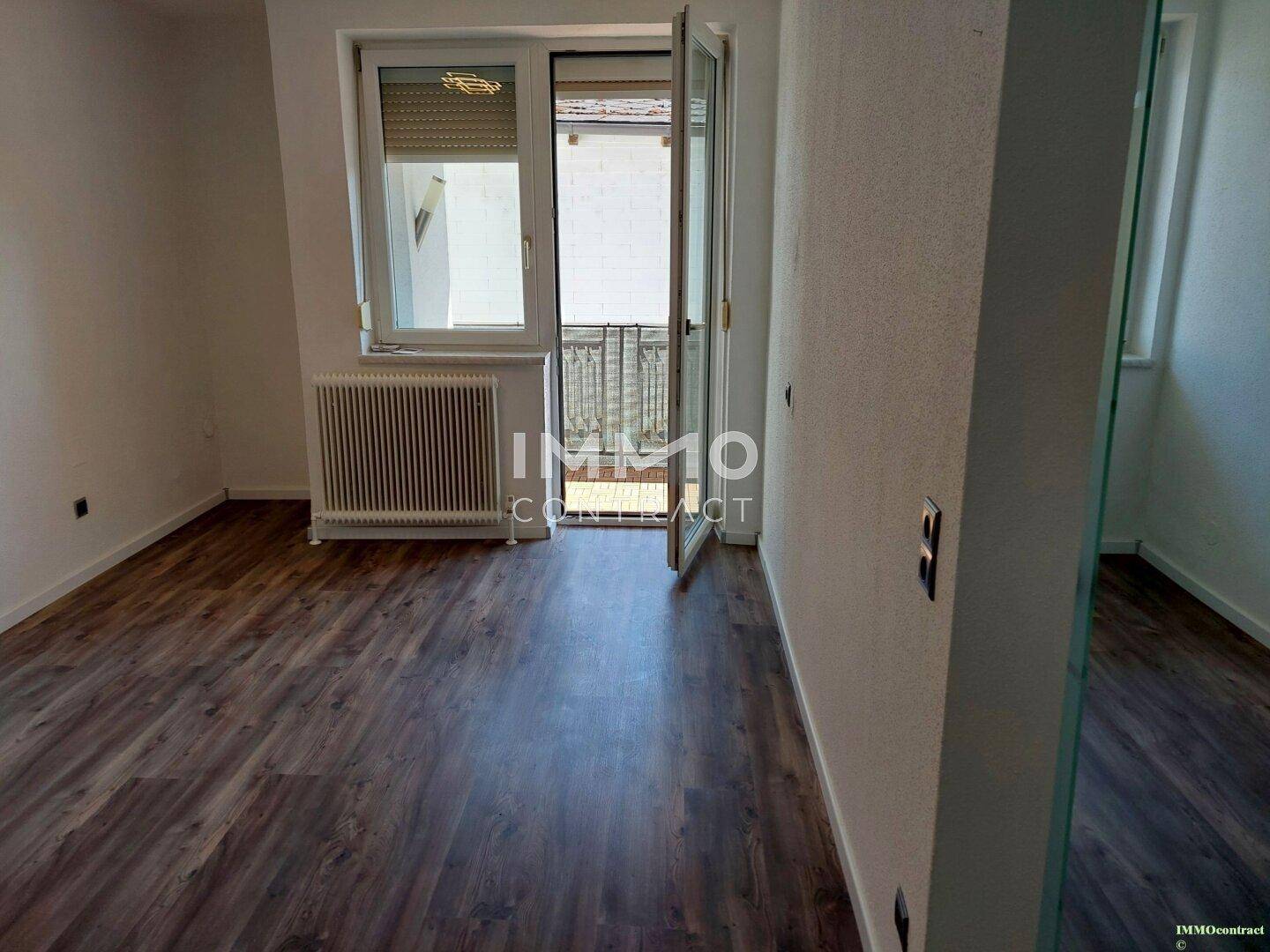 Heller Raum mit Laminatboden, Heizkörper und Zugang zu einem kleinen Balkon.