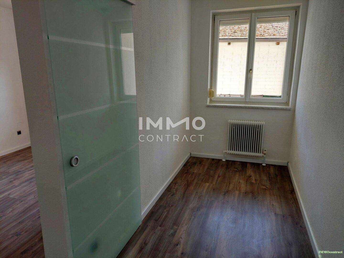 Kleines Zimmer mit Laminatboden, Fenster und einer modernen Glasschiebetür.