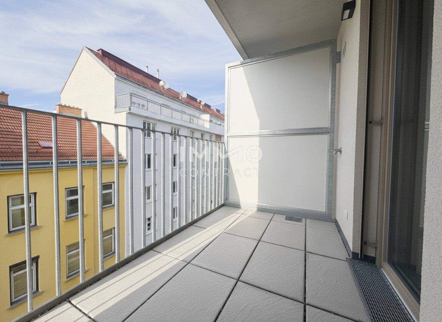 Heller Balkon mit Fliesenboden und Metallgeländer, bietet Blick auf umliegende Gebäude.