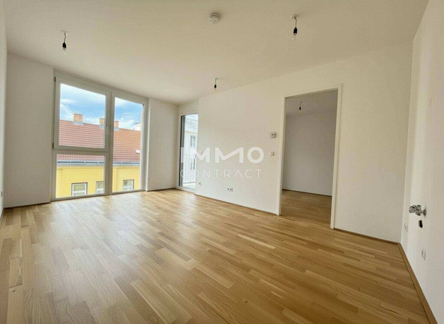 Heller, leerer Wohnraum mit großen Fenstern, Parkettboden und Zugang zu einem weiteren Zimmer.
