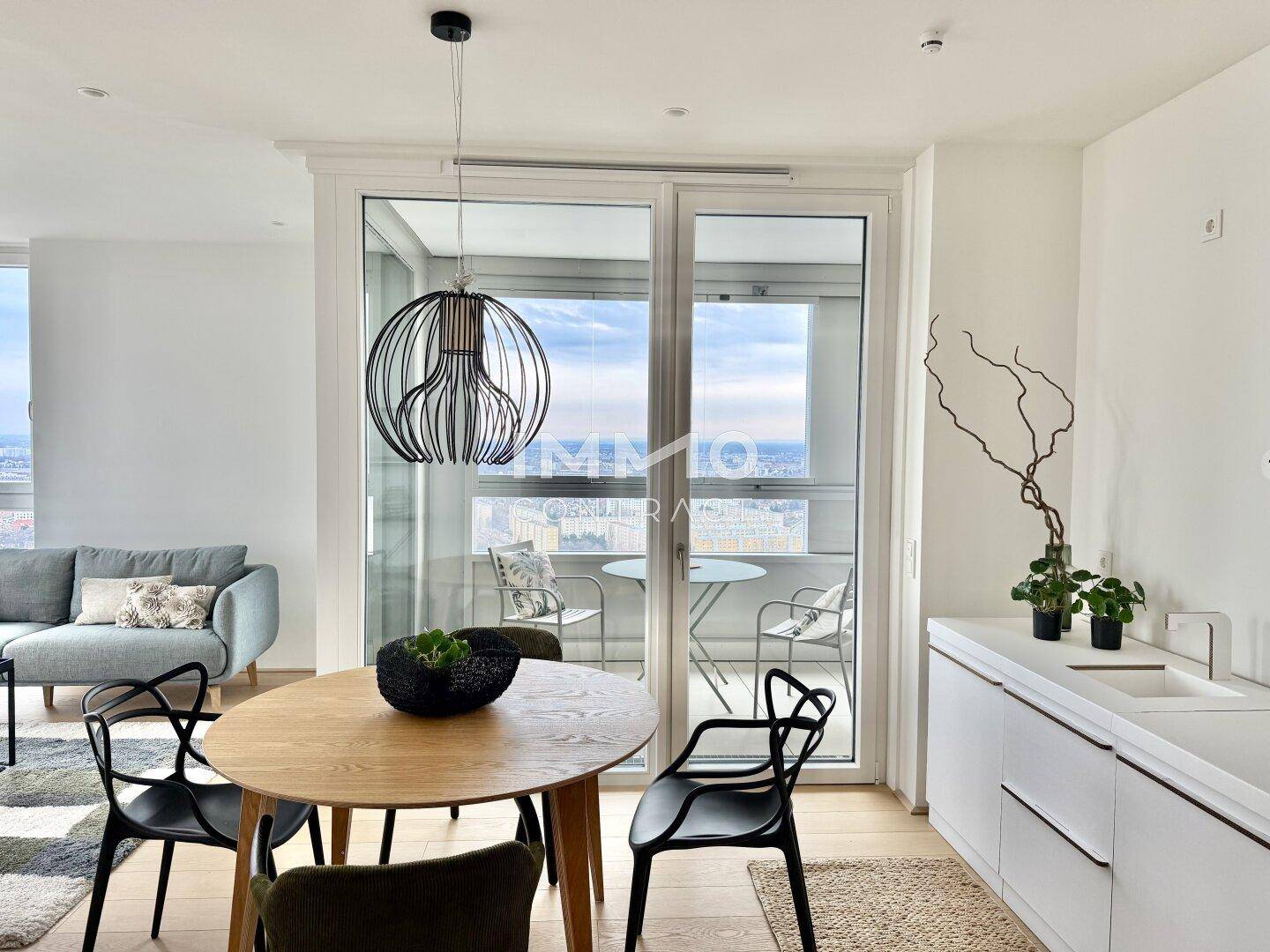 Moderner Essbereich mit Holztisch, minimalistischer Küchenzeile und Zugang zu einem Balkon mit Stadtblick.