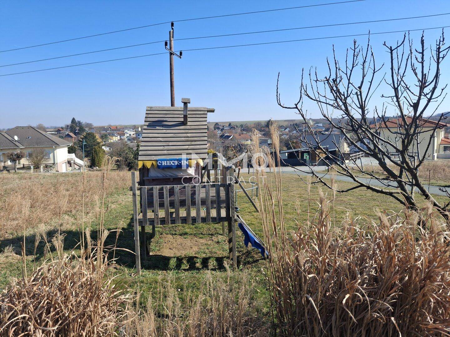 Hölzernes Spielhaus mit Rutsche im Garten, umgeben von hoher Vegetation.