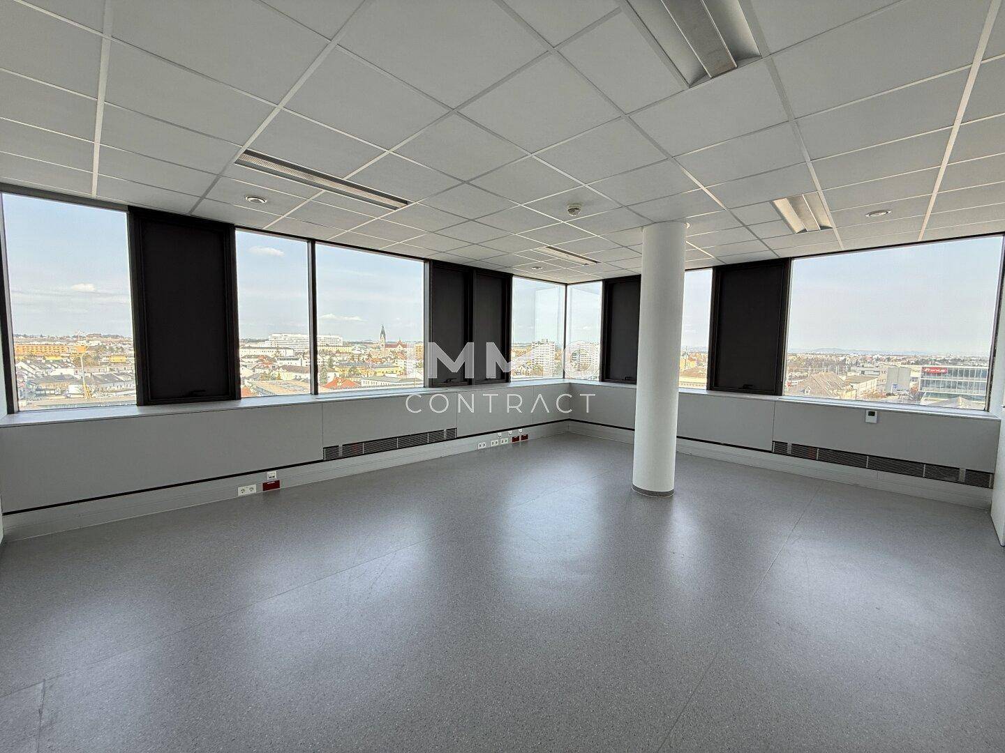 Leerer Büroraum mit hellem Vinylboden und großen Fenstern, die einen weiten Blick bieten.