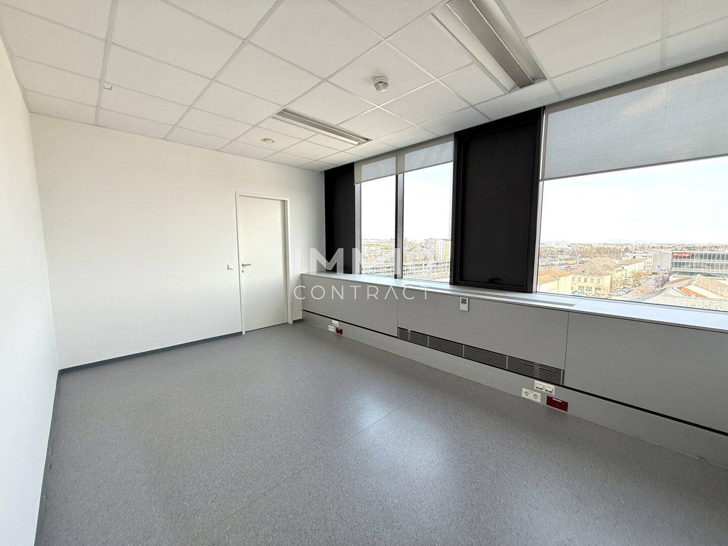 Kleinerer Büroraum oder Flur mit hellem Vinylboden, weißer Tür und großen Fenstern mit Stadtblick.