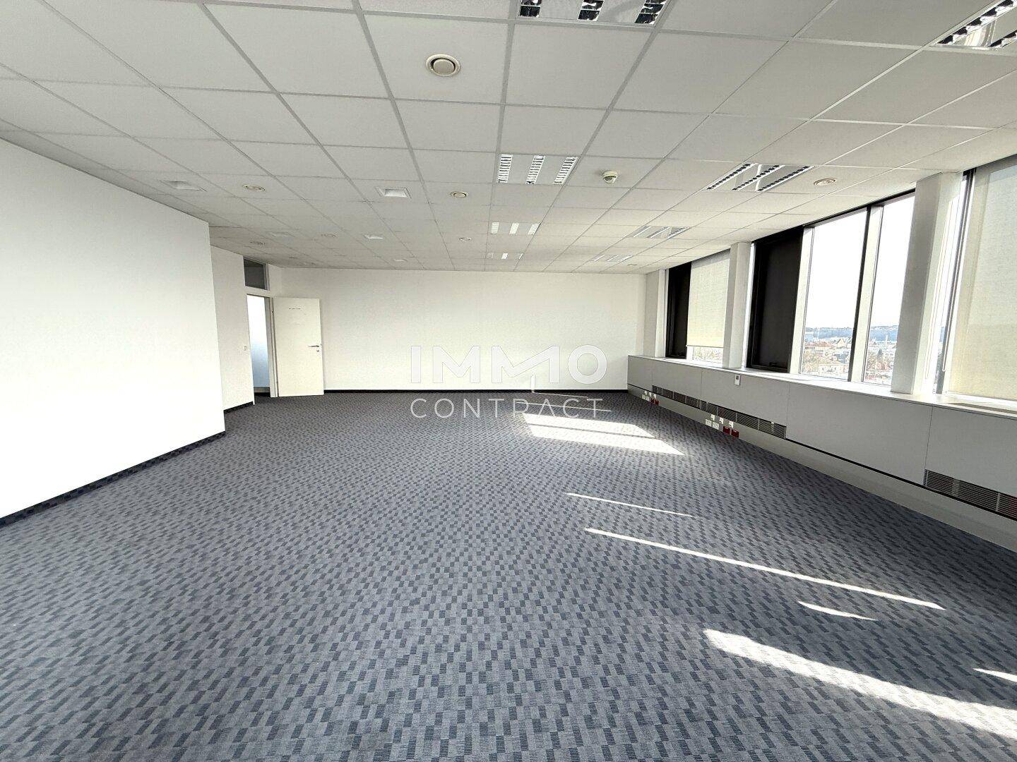 Großer, leerer Büroraum mit Teppichboden und Fenstern, die einen weiten Blick über die Stadt ermöglichen.