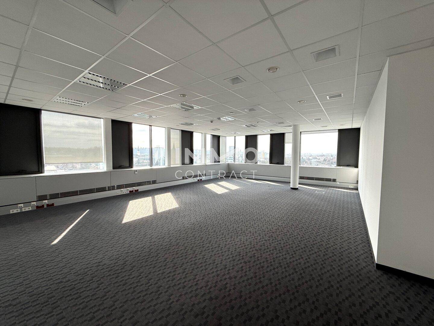 Geräumiger, leerer Bürobereich mit Teppichboden und umlaufenden Fenstern, die eine helle Atmosphäre schaffen.