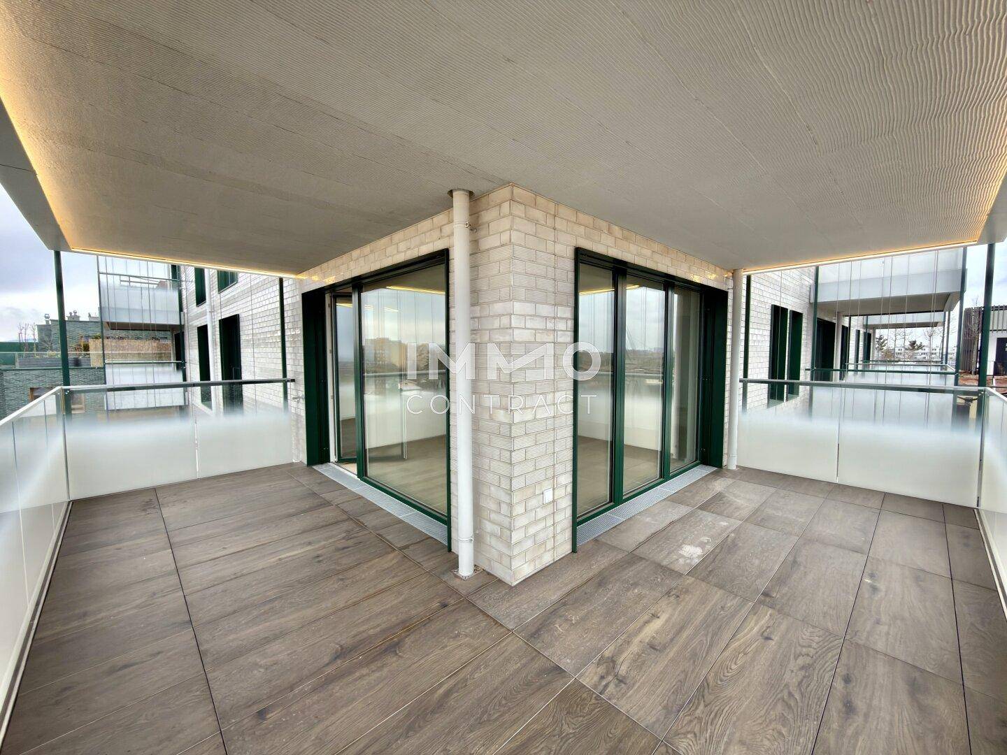 Großer Eckbalkon mit Fliesenboden und Glasgeländer, umgeben von moderner Architektur.