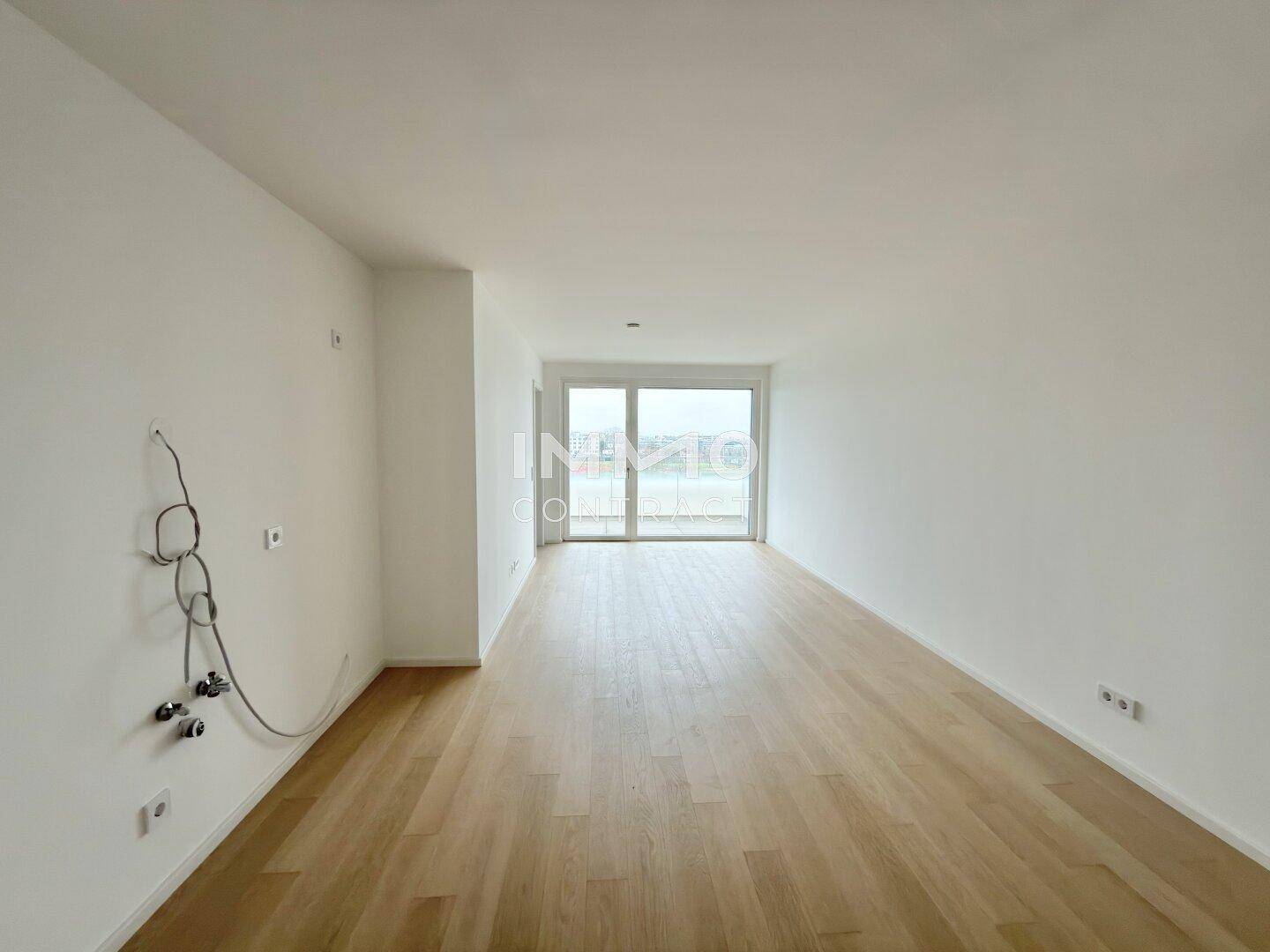 Helles, leeres Wohnzimmer mit Parkettboden und Zugang zu einem Balkon.