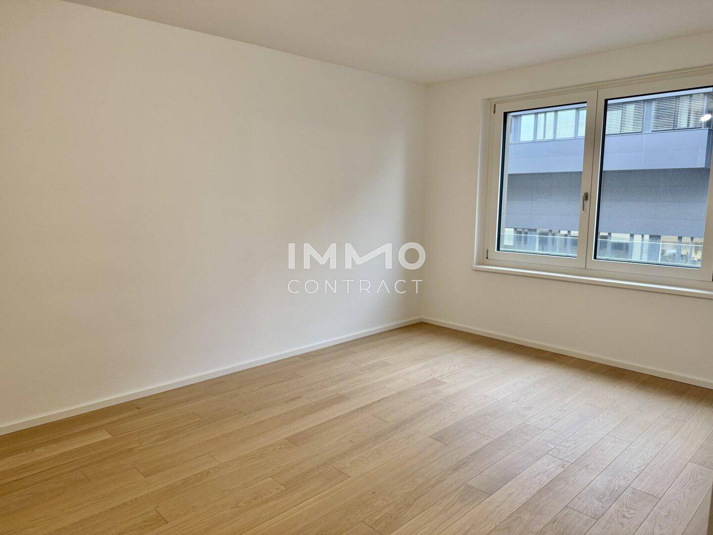 Geräumiges Zimmer mit hellem Parkettboden und Blick auf die umliegenden Gebäude.