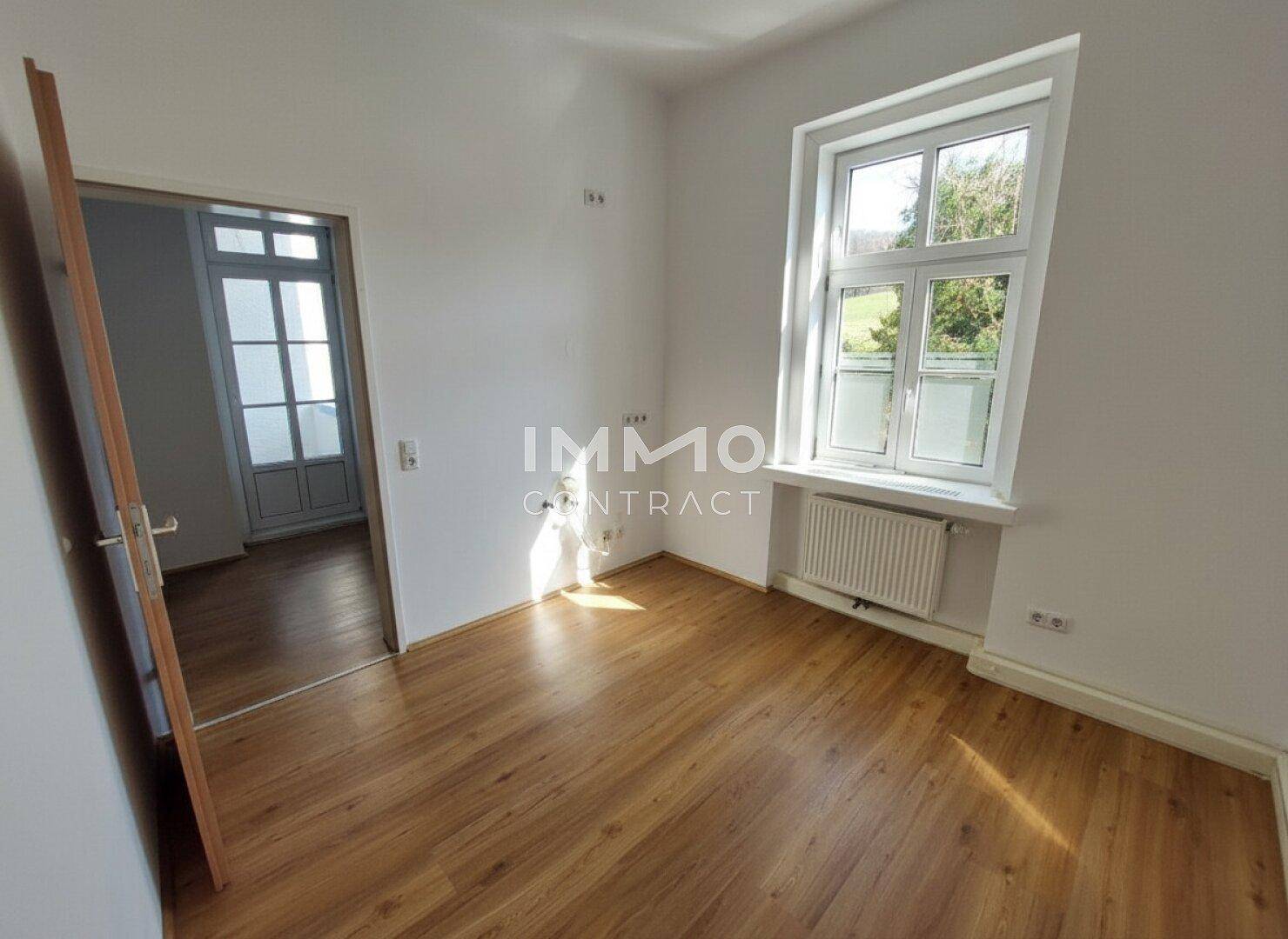 Zimmer mit Laminatboden, großem Fenster und Ausblick ins Grüne.