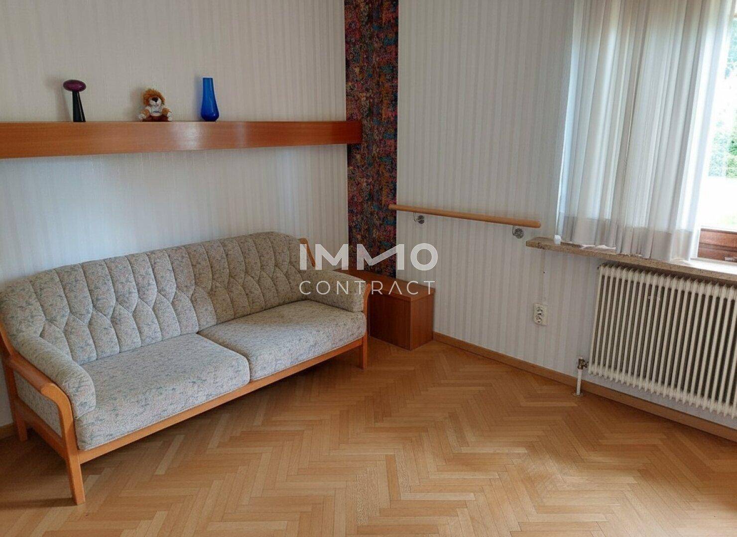 Helles Wohnzimmer mit Fischgrätparkett, bequemer Couch und Fenster mit Blick ins Grüne.