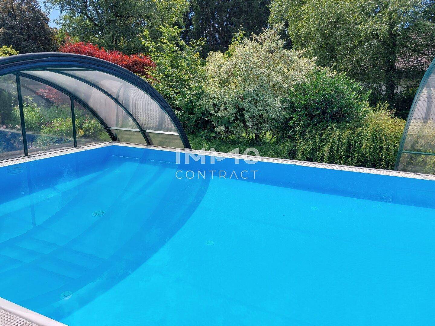 Blauer Swimmingpool mit Schiebeüberdachung, umgeben von üppigem Grün und Bäumen.