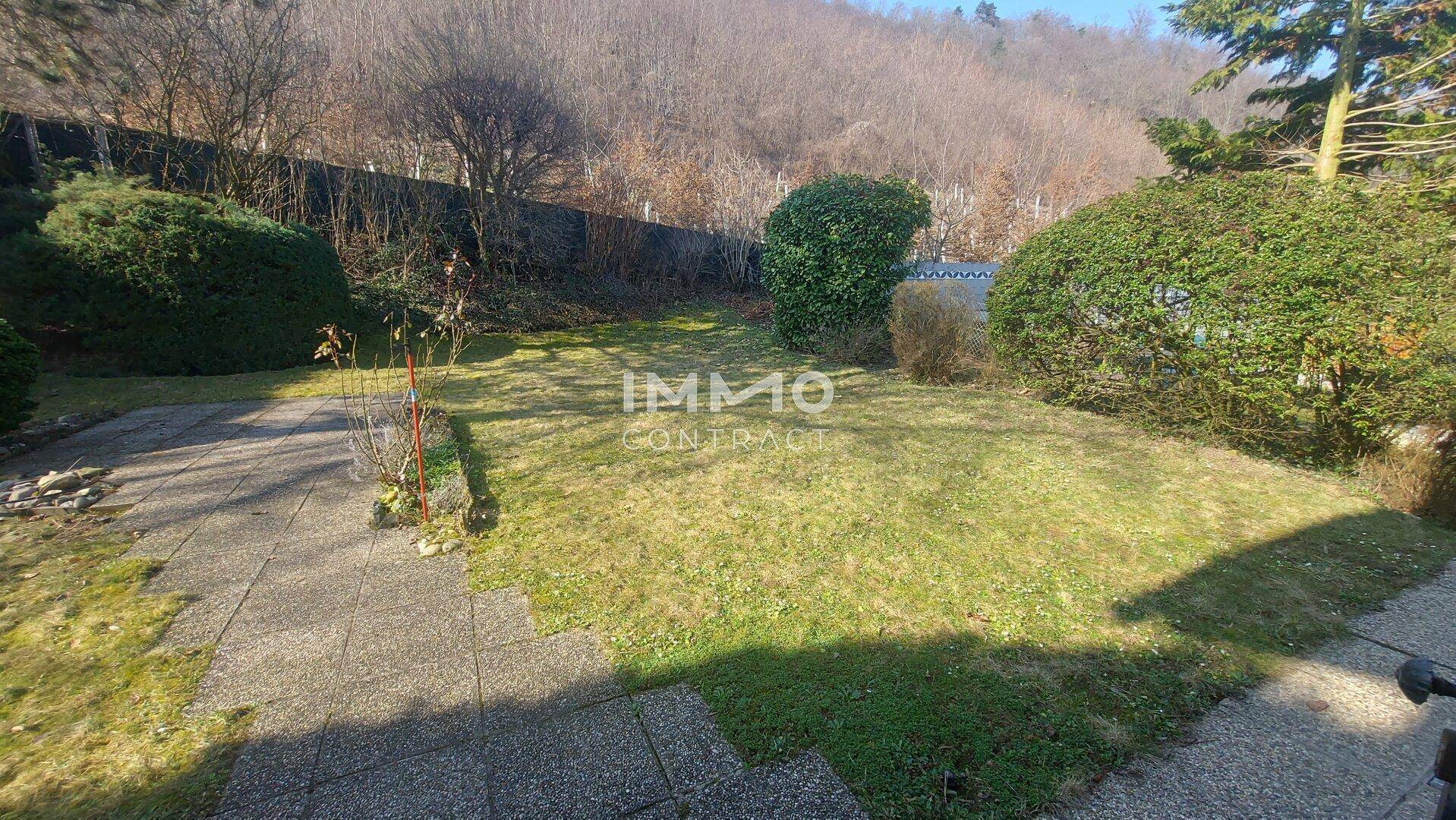 Gepflegter Gartenbereich mit Rasenfläche, Sträuchern und einem gepflasterten Weg, umgeben von Natur.