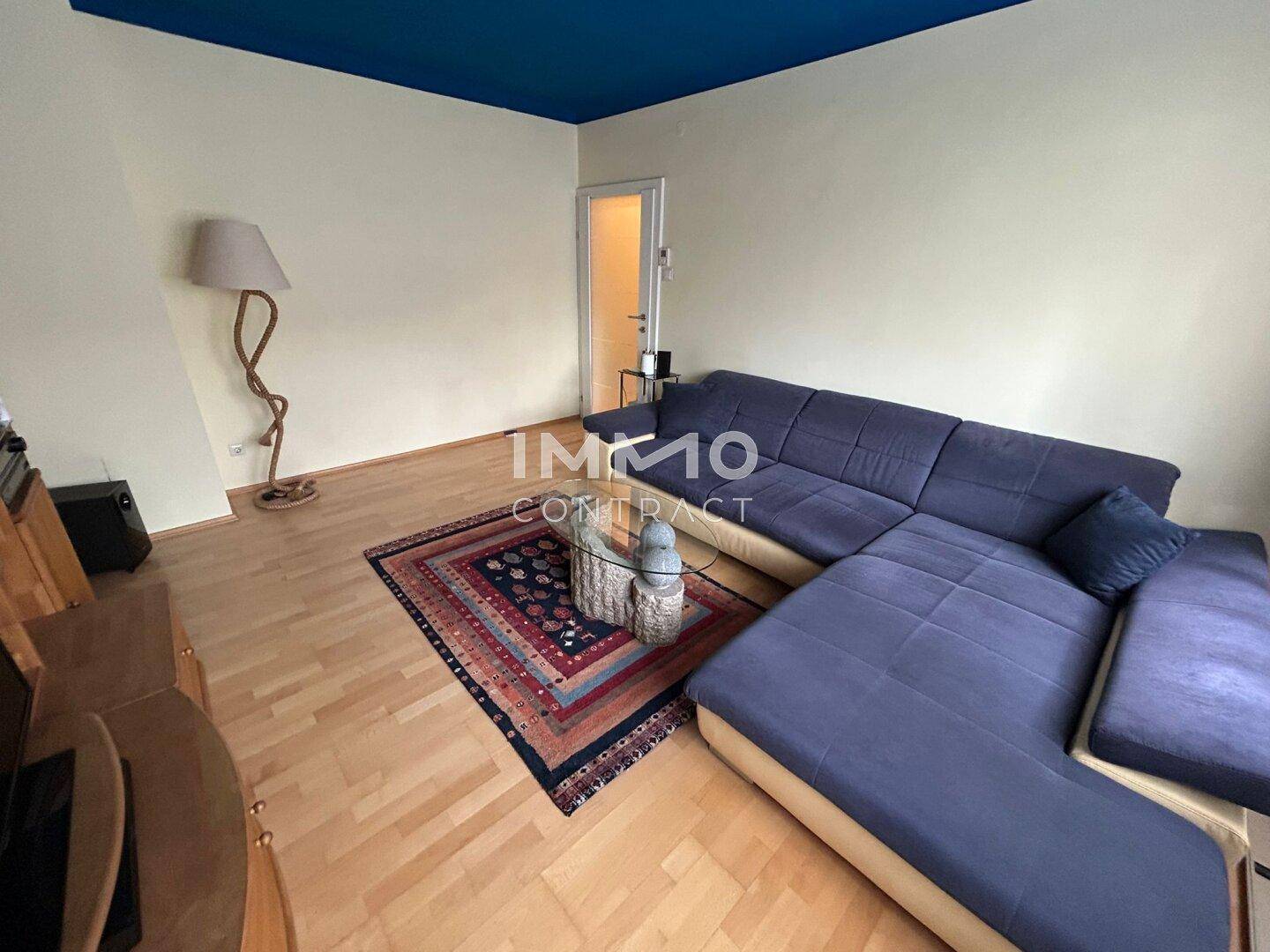 Wohnzimmer mit Ecksofa, Parkettboden und einer Tür, die zu einem weiteren Raum führt.