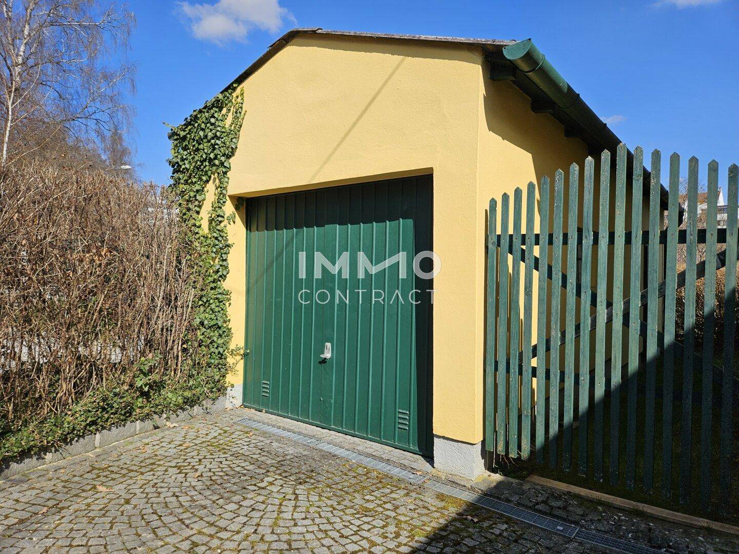 Einzelgarage mit grüner Tür und bewachsenen Wänden, umgeben von einem Zaun.